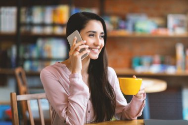 Kafede tek başına dinlenirken cep telefonuyla konuşan gülümseyen, gülümseyen ve gülümseyen Latin bayan. Mutlu Arap kadın akıllı telefonda konuşuyor ve çay ya da kahve içiyor.