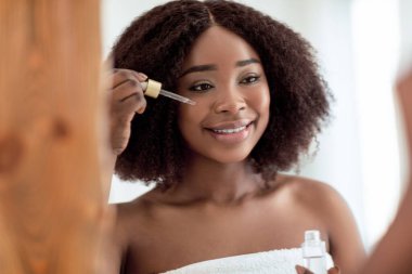 Evdeki aynanın yanında yüz serumu kullanan banyo havlusu giymiş neşeli siyah bir bayan. Gülümseyen Afrikalı Amerikalı kadın cildini nemli tutuyor, hyaluronik asit uyguluyor. Cilt bakımı ve sağlık konsepti