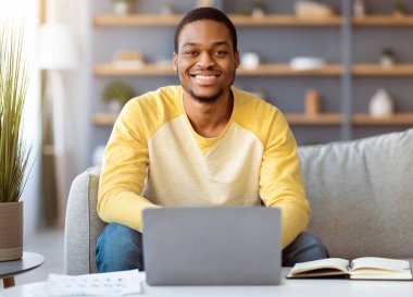 Evinden çalışan, kanepeye oturup dizüstü bilgisayar kullanan, not defteri olan serbest çalışan bir zenci. Neşeli Afrikalı Amerikalı genç iş adamı iş gününü planlıyor.