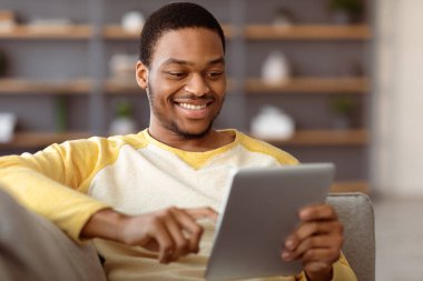 Dijital tablet ve iç mekan kullanarak kanepeye yaslanan pozitif siyahi adamın yakın çekimi. Mutlu Afrikalı Amerikalı adam oturma odasındaki koltukta dinleniyor, modern bloknotlardaki mobil uygulamaları kullanıyor, uzayı kopyalıyor.