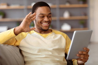 Neşeli Afrikalı Amerikalı genç adam arkadaşlarıyla veya sevgilileriyle video konferansı yapıyor, dijital tablet kullanıyor, tablet ekranlara bakıyor, gülümsüyor ve el sallıyor, ev içi, kopyalama alanı, yakın çekim