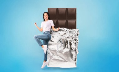 Neşeli bir kadın kolları havada, büyük bir çikolata parçasının yanında poz veriyor. Arkaplan canlı bir mavidir, o anın eğlenceli atmosferini arttırır..