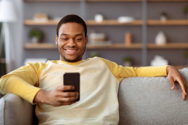 Rahat siyah adam evde tek başına kanepede oturuyor, akıllı telefon kullanıyor, arkadaşlarıyla sohbet ediyor, fotokopi çekiyor. Mutlu Afro-Amerikan genç adam internet sörfü yapıyor veya cep telefonunda sosyal medya kullanıyor.
