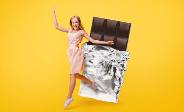 Çizgili elbise giyen neşeli bir kadın, parlak folyoya sarılı büyük bir çikolata çubuğunun yanında heyecanla zıplıyor. Parlak sarı arkaplan neşeli atmosfere ekleniyor.