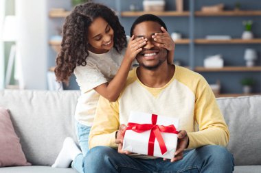 Babalar Günü konsepti, kıvırcık saçlı tatlı Afro-Amerikan kızı babasını selamlıyor, kızı mutlu babasının gözlerini kapıyor ve ona hediye kutusu veriyor, doğum gününü evde kutluyor, boşluğu kopyalıyor.