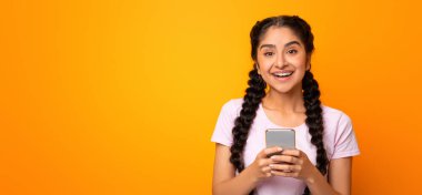 Harika Uygulama Konsepti. Sarı stüdyo arka planında kameraya bakarak izole edilmiş akıllı telefon kullanan mutlu, genç Hintli kadın portresi. İnternette sohbet eden kadın, sosyal medyada gezinen.