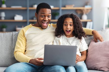 Oturma odasındaki kanepede otururken bilgisayar kullanan siyah baba ve kızı şok oldu. Heyecanlı Afro-Amerikan ailesi evde dizüstü bilgisayar kullanıyor, heyecan verici bir şey izliyor, uzayı kopyalıyor.