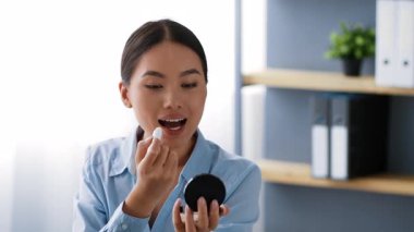 Açık mavi gömlekli genç bir Asyalı kadın büyük bir pencerenin önünde masasında oturuyor. Ruj sürerken aynasını kullanıyor ve modern bir ofiste iş gününe hazırlanıyor..