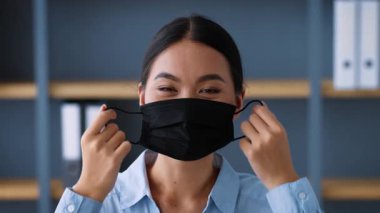 Genç bir Asyalı kadın çalışma alanında çalışırken siyah bir maske takıyor. Profesyonel bir ortamda güvenliğin önemini vurgulayarak görevlerine odaklanmış durumda..