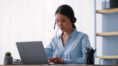 Genç bir Asyalı kadın, parlak ve modern bir ofis alanında kulaklık takarken dizüstü bilgisayarına odaklanıyor. İşiyle meşgul, profesyonelliğini ve konsantrasyonunu gösteriyor..