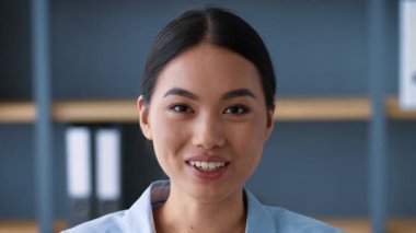 Profesyonel olarak giyinmiş genç bir Asyalı kadın modern ofis ortamında kameraya sıcak bir şekilde gülümsüyor. İşbirlikçi bir ortamda çalışırken kendinden emin ve meşgul görünüyor..