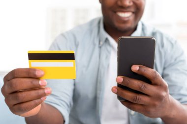 Banka işlemleri. Kredi kartı tutan ve evden alışveriş yapmak için akıllı telefon kullanan mutlu Afro-Amerikalı adam. Para çekmeceleri için mobil uygulama, fotokopi alanı. E-ticaret kavramı