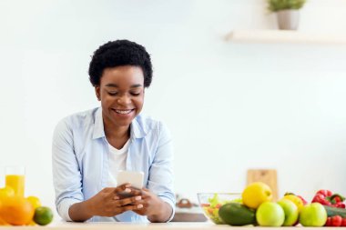 Siyah kadın, yemek pişirme uygulaması olan telefonu kullanıyor. Sağlıklı yemek hazırlıyor ve evde modern mutfakta yemek tariflerini tarıyor. Beslenme Uygulaması Konsepti. Boşluğu Kopyala