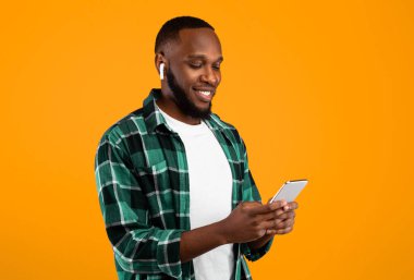 Music Application. African Man Using Phone Wearing Earbuds And Listening To Song Online Standing Over Yellow Studio Background. Smartphone User Browsing Internet On Cellphone