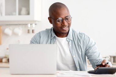 Faturaları ödemek, internette vergi ödemek ve aile bütçesini hesaplamak. Olgun Afro-Amerikalı adam mutfakta evrak ve dizüstü bilgisayarla çalışıyor.