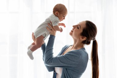 Mutlu genç anne, yeni doğmuş bebeğini kaldırırken pencerenin yanında duruyor, milenyum annesini seviyor, bebekle oynuyor, onu tutuyor ve gülümsüyor, boşluğu kopyalıyor.