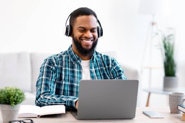 Bilgisayarda çalışan ve kulaklık takan mutlu bir Afrikalı adam modern ofiste işyerinde oturuyor. İş ve Girişimcilik, Başarılı Kariyer ve Mesafe Konsepti. Seçici Odaklanma