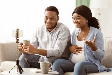 Neşeli siyahi adam ve kadın blogcular bekliyor, video blogcular evden yayın yapıyor, yaşam tarzlarını takipçilerle paylaşıyorlar, akıllı telefonu tripodlarda kullanıyorlar, oturma odası içlerinde, hamilelik blogu konseptinde