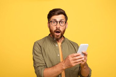 Sakallı orta yaşlı bir adam, yeşil gömlek giymiş akıllı telefonuna bakarken şaşırdığını ifade eder. Parlak sarı bir arka plana karşı duruyor ve neşeli bir atmosfer yaratıyor..