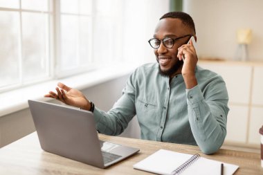 Modern Ofis 'te iş yerindeki dizüstü bilgisayarın başında cep telefonuyla konuşan olumlu siyahi işadamı. Çalışanların iş yerindeki cep telefonuyla konuşmaları. İş İletişimi