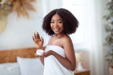 Genç cildiyle ilgilenmek. Duştan sonra yüz kremi süren, içeride kameraya gülümseyen duygusal siyahi kadın. Afro-Amerikalı kadın evde güzellik tedavisi uyguluyor.