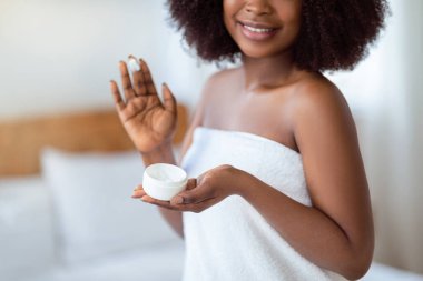 Cilt bakımı, kaplıca salonu. Çekici, banyo havlusuyla genç Afrikalı Amerikalı kadın. Yüzüne doğal nemlendirici krem sürüyor. Yakın çekim görüntüsü.