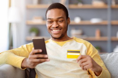 Kredi kartı ve cep telefonu olan neşeli Afrikalı Amerikalı genç adam evdeki kanepede oturuyor, yakın plan, kopyalama alanı. İnternetten alışveriş yapan mutlu siyahi adam, akıllı telefon ve kartvizit kullanıyor, e-ticaret yapıyor.