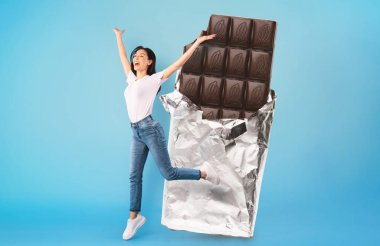 Bir kadın folyoya sarılı kocaman bir çikolatanın yanında neşeyle dans ediyor. Parlak mavi arka plan, o anın neşeli atmosferini arttırır..