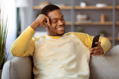 Neşeli Afrikalı Amerikalı genç adam evde tek başına takılıyor. Yepyeni bir akıllı telefon kullanıyor. Kopya alanı. Oturma odasındaki koltukta oturan mutlu siyah adam cep telefonunda yeni bir flört uygulaması kullanıyor.