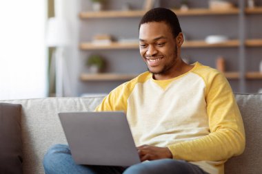 Gülümseyen Afrikalı Amerikalı genç adam Webinar 'a katılıyor, evinde dizüstü bilgisayar kullanıyor, uzayı kopyalıyor. Neşeli siyahi adam kız arkadaşıyla online sohbet ediyor, online arkadaşlık sitelerini ziyaret ediyor.