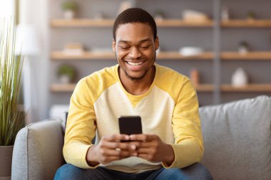 Evdeki kanepede oturan, akıllı telefon kullanan, arkadaşlarıyla sohbet eden, internette oynayan, kopya alan gülümseyen siyah adam. Mutlu Afro-Amerikan genç adam internet sörfü yapıyor veya cep telefonunda sosyal medya kullanıyor.
