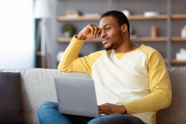 Evinden online çalışan hayalperest siyahi adam, modern dizüstü bilgisayarla koltukta oturuyor, fotokopi alanına bakıyor ve gülümsüyor. Yakışıklı Afro-Amerikan adam internetten iş arıyor.