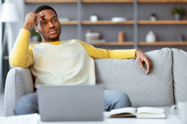 Düşünceli genç siyahi iş adamı dizüstü bilgisayarın önünde oturuyor, kafasına dokunuyor ve fotokopi alanına bakıyor, salgın hastalık salgını, iç mekan