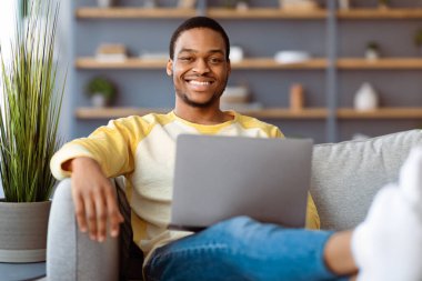 Kanepede keyfi takılan, dizüstü bilgisayar ve iç mekan kullanan pozitif siyah bir adam. Mutlu Afro-Amerikalı adam oturma odasında dinleniyor, sosyal medyada sörf yapıyor, online randevusu var, uzay kopyası yapıyor.