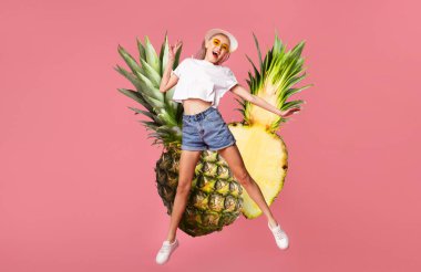 Beyaz bir bluz ve güneş gözlüğü takan genç bir kadın iki büyük ananas arasında heyecanla zıplıyor. Parlak pembe arka plan, o anın canlı atmosferine ekleniyor..