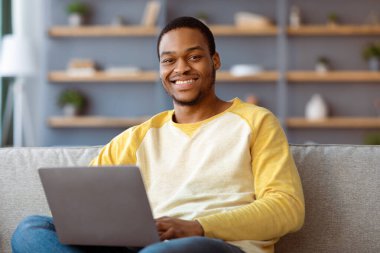 Modern dizüstü bilgisayarı olan neşeli siyah adam evdeki kanepede dinleniyor, kameraya bakıyor ve gülümsüyor. Mutlu Afrikalı Amerikalı genç adam defter kullanıyor, sosyal medyada sörf yapıyor, iç mekanda oturuyor, yakın çekim yapıyor.