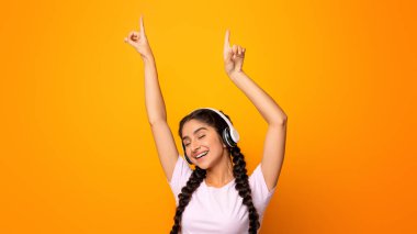 Favori şarkının tadını çıkarıyorum. Duygusal heyecanlı Hintli kadın kablosuz kulaklık takıyor müzik dinliyor, gözleri kapalı dans ediyor ve ellerini kaldırıyor. Pozitif kadın eğleniyor, sarı stüdyo duvarı