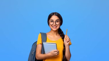 Cevabı biliyorum. Güzel gülümseyen gözlüklü Hintli kız öğrencinin portresi. Elinde not defteri, elinde kalemle mor stüdyo arka planına izole edilmiş sırt çantası.