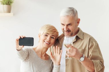 Tatilde karşılama, evde kutlama yapan bir çift ve video görüşmesi. Elinde beyaz şarap kadehi olan gülümseyen yetişkin bir adam, akıllı telefonlu bir bayan ve modern mutfağın içinde webcam 'e el sallıyor.