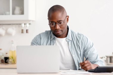 Bütçe planlama konsepti. Düşünceli Afro-Amerikan adam vergileri hesaplıyor, dizüstü bilgisayar kullanıyor, evde mutfakta oturuyor. Tasarruf, finans ve ekonomi kavramı.