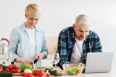 Mutlu orta yaşlı çift dizüstü bilgisayar kullanıyor, sağlıklı yemek, diyet, evde sebze salatası hazırlıyor, yemek tariflerini arıyor, internetten alışveriş yapıyor, mutfakta yemek kurslarını izliyor, panorama