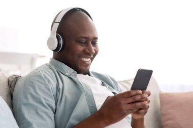 Rahat orta yaşlı Afro-Amerikalı adam akıllı telefonuyla müzik dinliyor, cep telefonuyla favori şarkılarının tadını çıkarıyor, oturma odasında rahat bir koltukta dinleniyor, boş yer var.