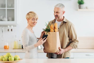 Modern alışveriş, nakit para ve sipariş ürünleri, eve teslimat. Gülümseyen sarışın kadın mesaj yazıyor ya da telefona not veriyor ve kocası tarafından mutfağa getirilen erzak paketlerine bakıyor.