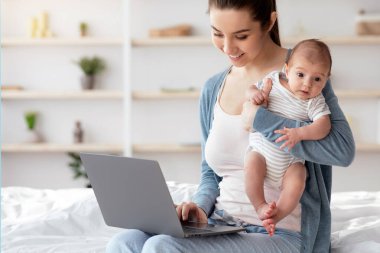Modern Annelik Konsepti. Dizüstü bilgisayarda çalışan ve kucağında yeni doğmuş bebeği tutan genç ve güzel bir kadın, Milenyum Serbest Kadın Uzaktan Çalışmayla Çocuk Bakımı 'nı birleştiren,