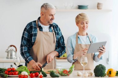 Evde modern teknolojilerle sağlıklı yemekler hazırlayın. Mutlu yetişkin adam sebze keser, kadın mutfak içinde tablete bakar masada parlak sebzeler ve şarap kadehleri, boş alan