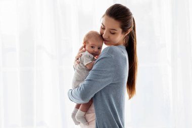 Güzel, genç bir anne, yeni doğmuş bebeğini kucağında tutarken pencerenin kenarında, sevgi dolu bir anne, bebeğini kucaklıyor, anneliğin tadını çıkarıyor, boşluğu kopyalıyor.