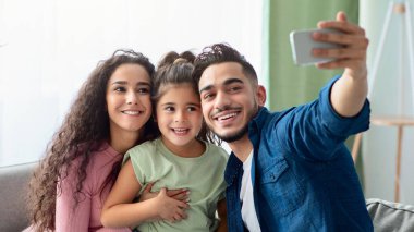 Üç kişilik mutlu bir Arap ailesi evde akıllı telefonlarıyla selfie çekiyor, neşeli orta doğulu ebeveynler ve tatlı küçük bir kız çocuğu oturma odasında birlikte rahatlarken fotoğraf çektiriyorlar.