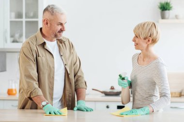 Birlikte temizlik yapmaktan hoşlanan mutlu bir çift, ev hijyeni, ilişkiler ve ev hayatı. Mutlu orta yaşlı karı koca koruyucu eldivenlerle mutfak içi dezenfekte masası, boş alan.