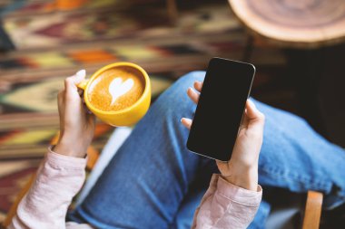 Kafeteryada kahve içerken siyah ekranlı telefon kullanan genç bir kadın. Tasarımın için akıllı telefon. Cep telefonunda mobil uygulama ya da web sitesi için boşluk tutan kadın