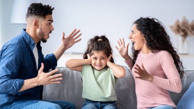 Ebeveyn çatışmaları. Elleriyle kulaklarını kapatan üzgün küçük Arap kız ebeveynlerin tartışmalarını dinlemiyor bağıran anne ve baba arasında koltukta oturan sinirli çocuk evde çıkan aile içi kavgalara tanık oluyor.
